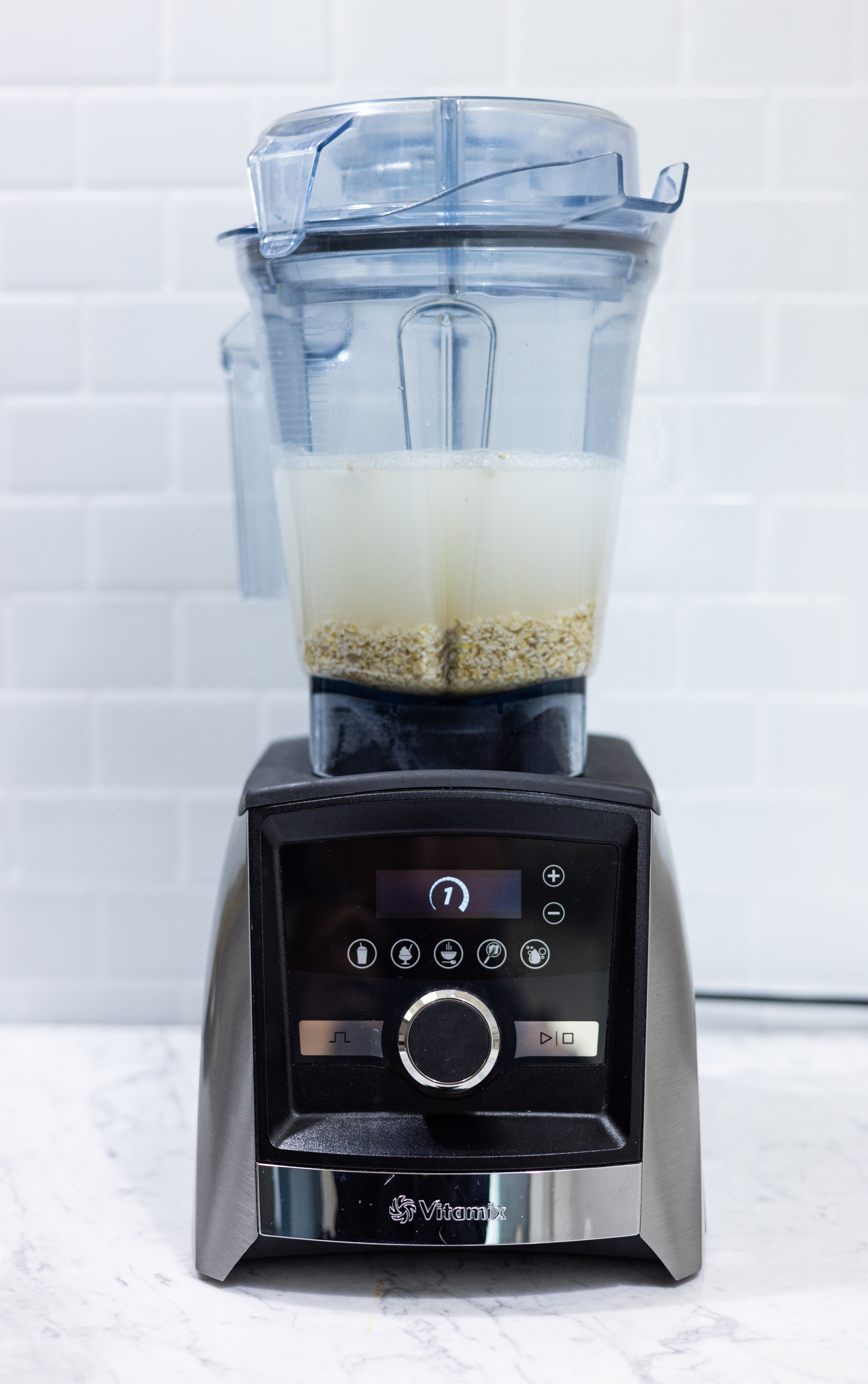 In this picture you can see a Vitamix A350i Ascent series a luxurious high-end blender and the best one of the market. The vitamix is filled with organic rolled oats and filtered organic water. The vitamix. The background is plastered with white metro tiles. The blender stands on carrara marble.