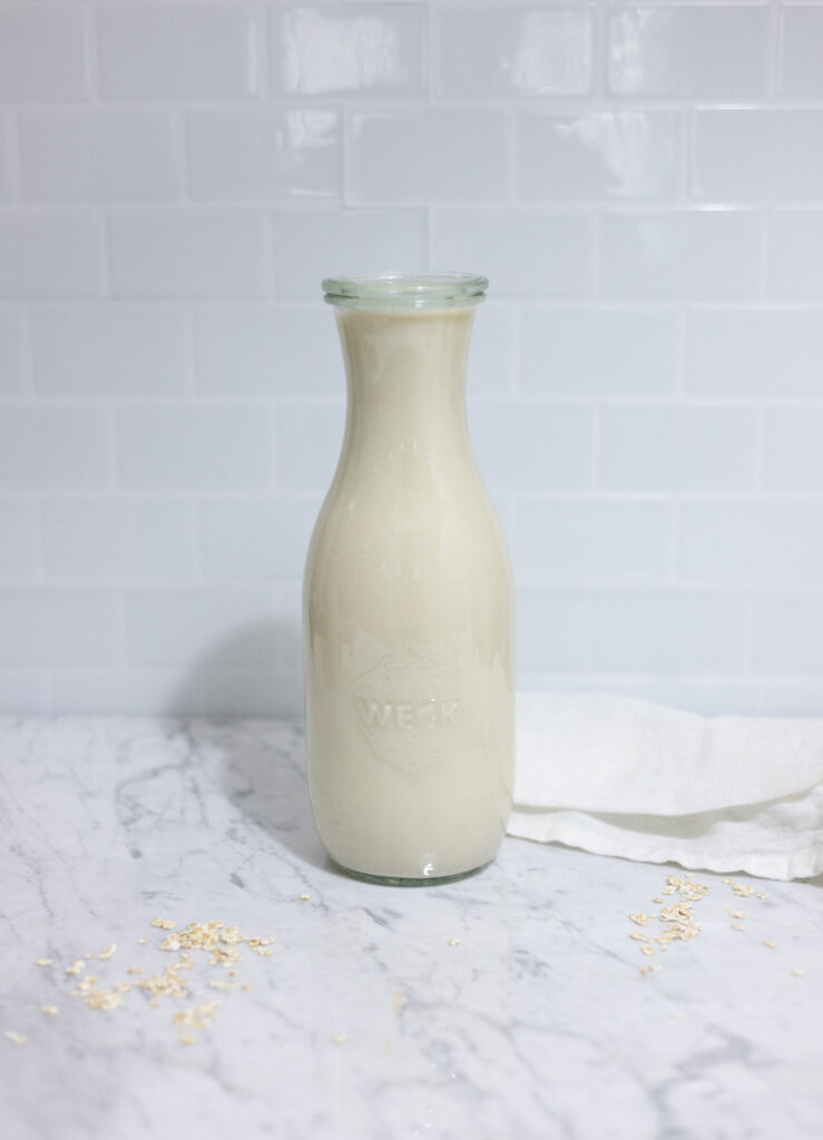 In this picture you can see a weck glass full of homemade oat milk creamer. The homemade oat milk creamer is plated on a marble desk with a white linen towel in the background. The background is plastered with white metro tiles.