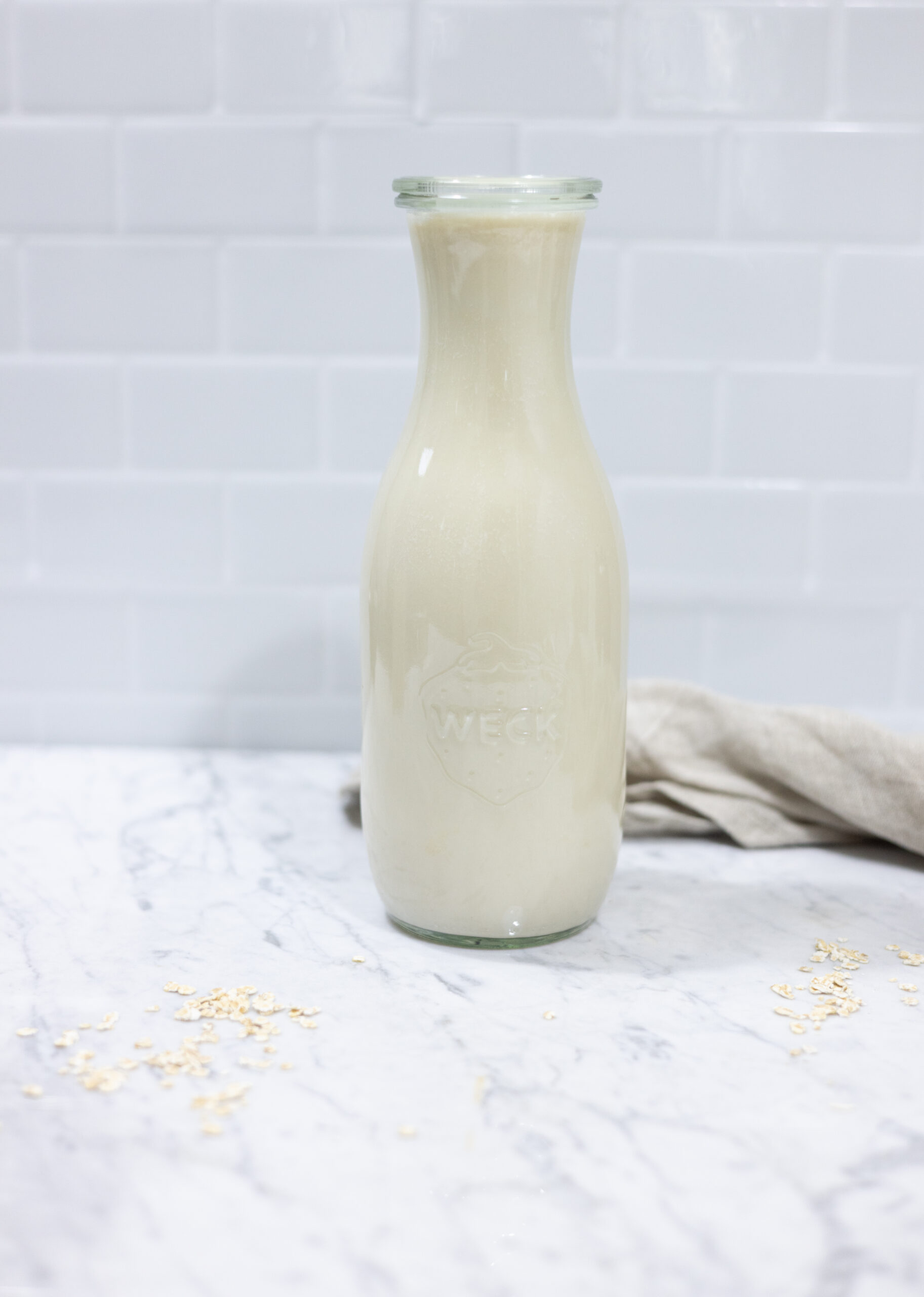 In this picture you can see a weck glass full of homemade oat milk creamer. The homemade oat milk creamer is plated on a marble desk with a beige linen towel in the background. The background is plastered with white metro tiles.