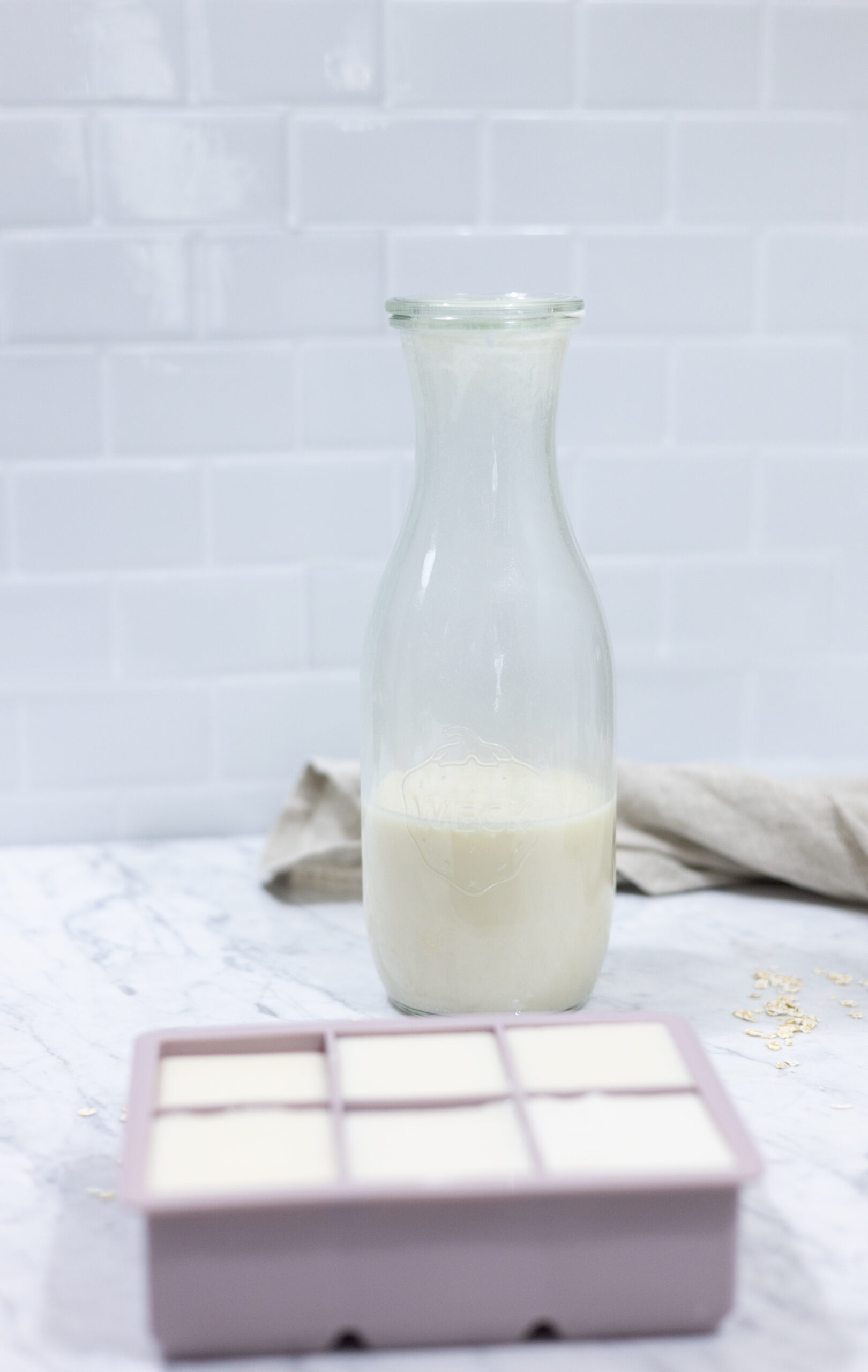 In this picture you can see a weck glass full of homemade oat milk creamer. The homemade oat milk creamer is plated on a marble desk with a beige linen towel in the background. In the front is an ice cube mold in which the homemade oat creamer is filled in. Besides it, you can see singular oats placed as a decoration. The background is plastered with white metro tiles.