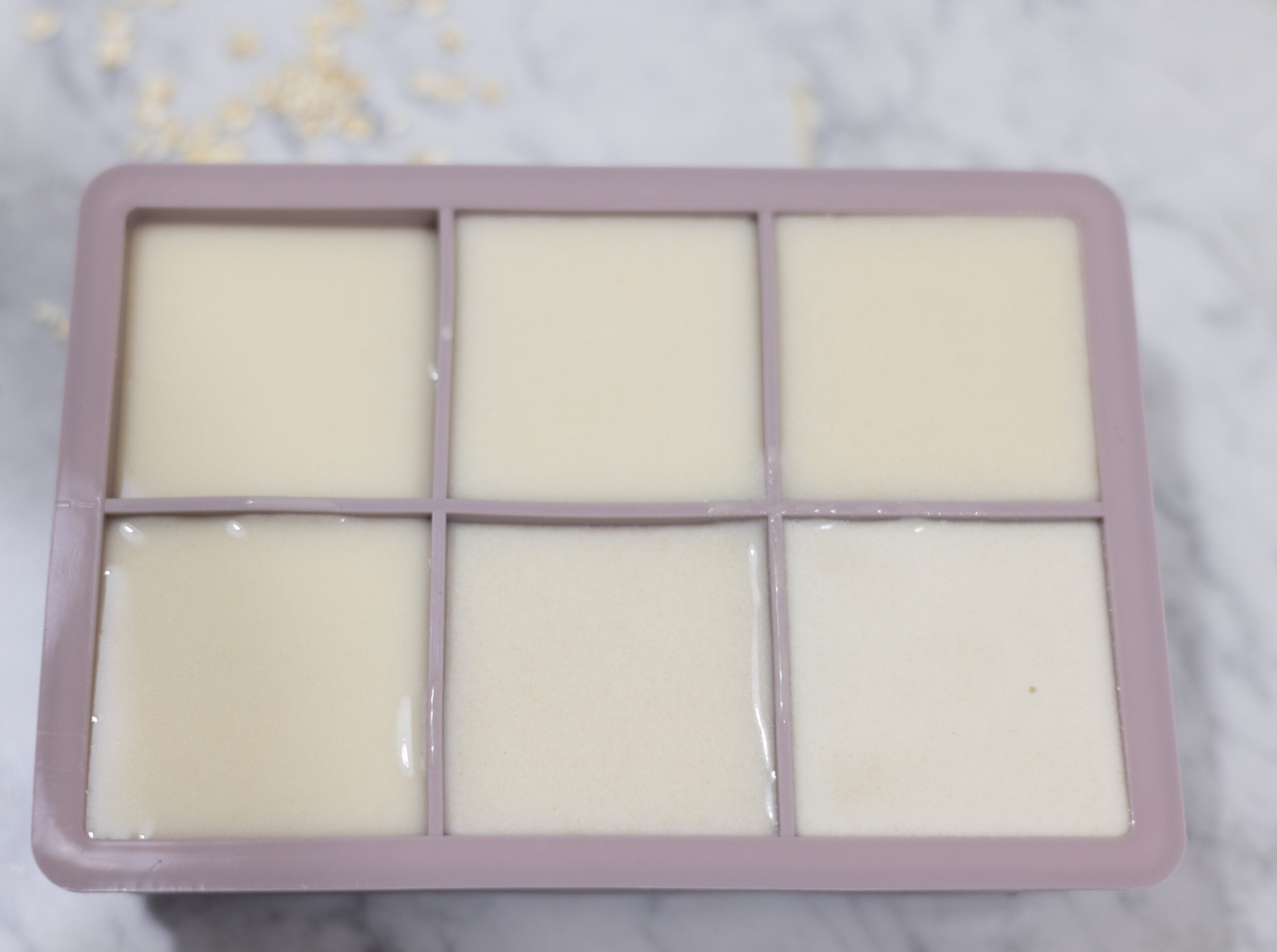 In this picture you can see the purple ice cube mold, in which the homemade oat creamer is filled in. The homemade oat milk creamer is plated on a marble desk with a beige linen towel in the background. In the front is an ice cube mold in which the homemade oat creamer is filled in. Besides it, you can see singular oats placed as a decoration. The background is plastered with white metro tiles.