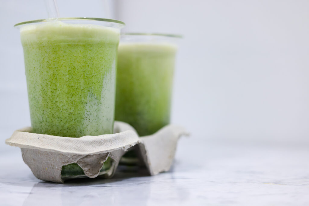 Here you can see the finished Iced Vanilla Matcha cups. These are supported by cardboard holders and stand on Carrara marble. The metro tiles can be seen in the background. There are glass straws in the cups for direct enjoyment.