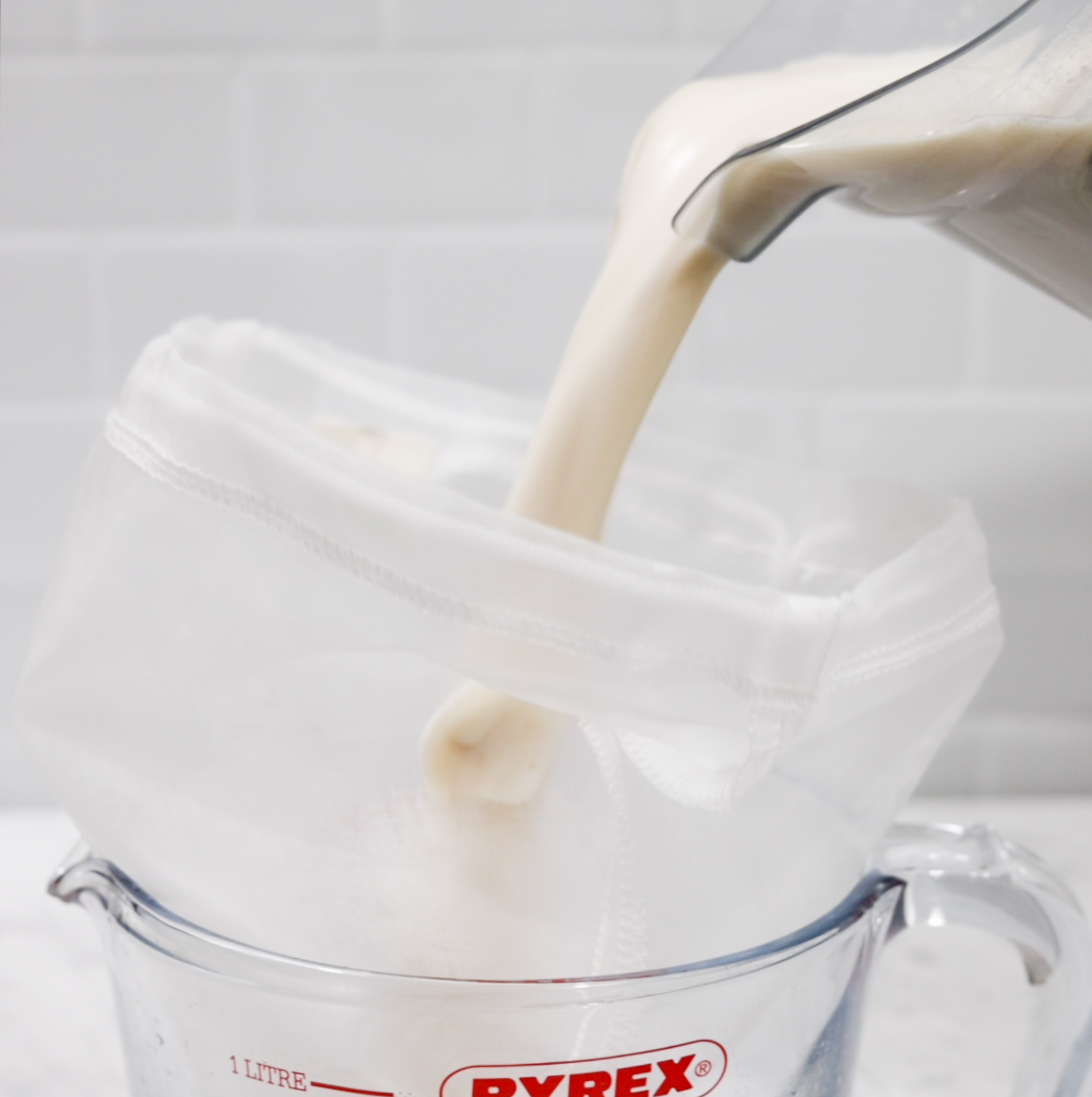 In this picture you can see a nut bag placed in a Pyrex glass jar.  Meanwhile it gets filled with the homemade oat milk creamer. The homemade oat milk creamer has a beige milky color. The background is plastered with white metro tiles. The blender stands on carrara marble.