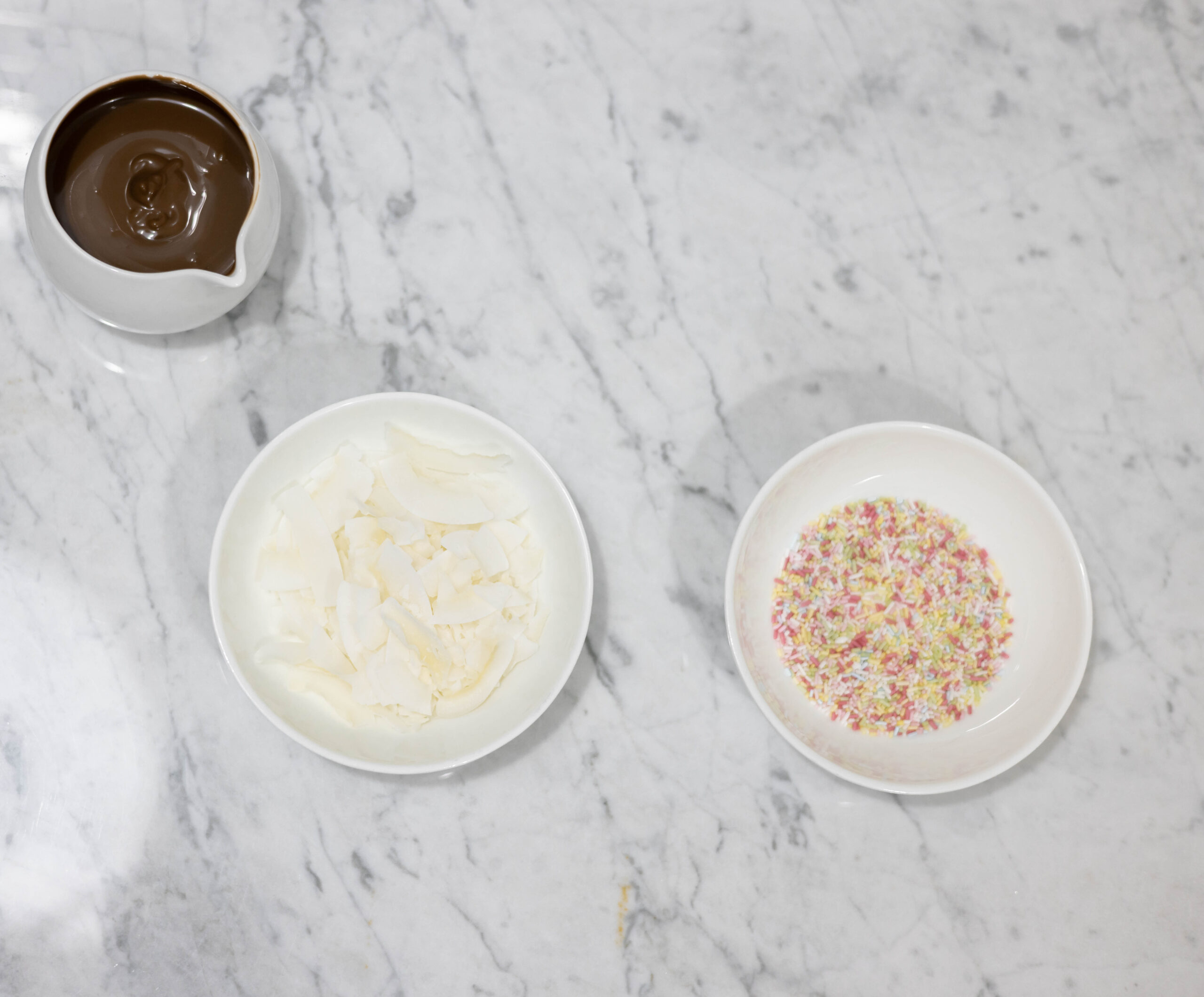 In this picture you can see a fine bone china jar filled with vegan warm chocolate. 
Next to the chocolate jar are two a small bowls made of fine bone china porcelain filled with a garnish ingredient named organic colorful sprinkles and shredded coconut. The whole setting is positioned on a white Carrara marble desk.
