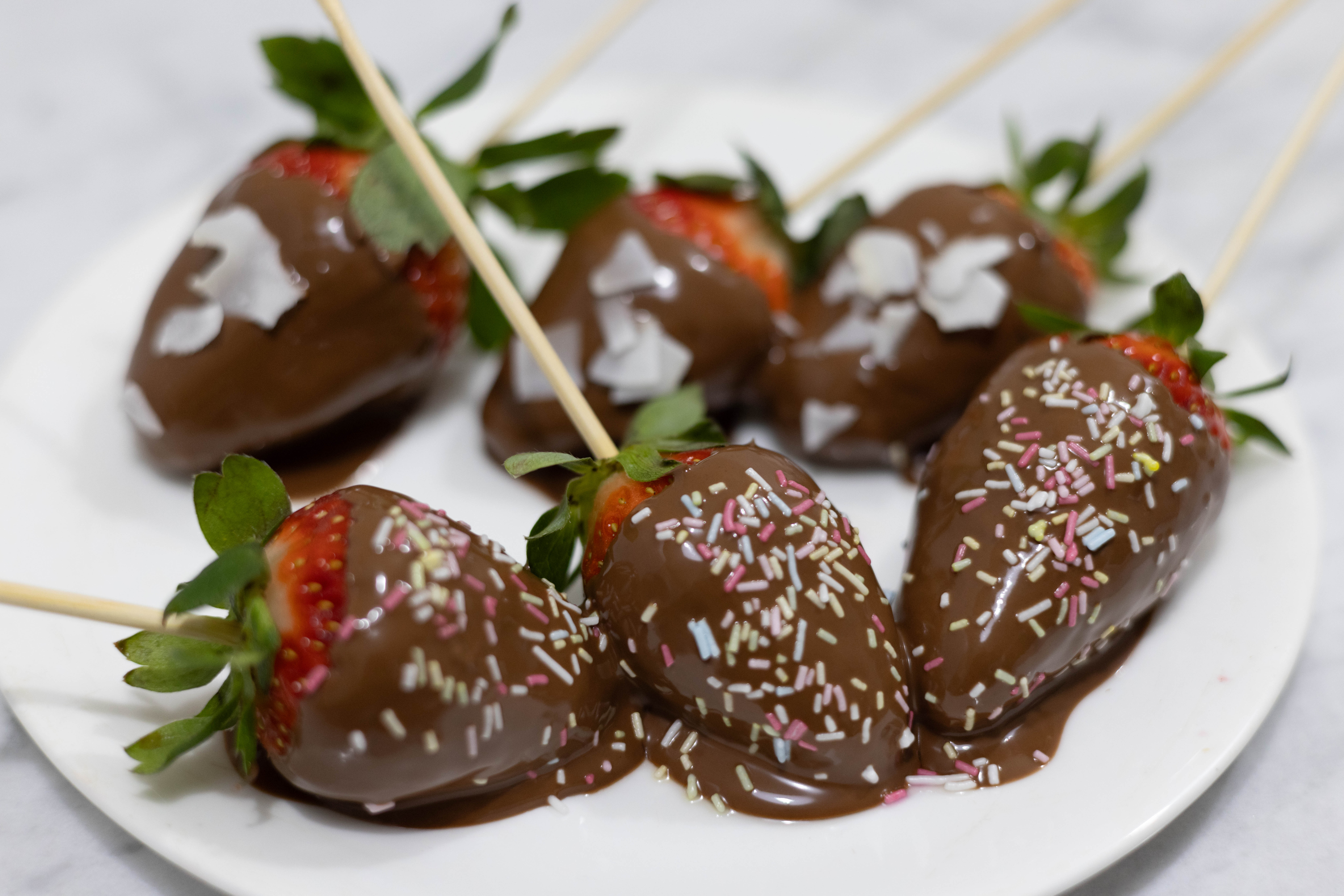 In this picture you can see the six Vegan Chocolate-Covered Strawberries plated. Three are garnished with organic colorful sprinkles and the other three a garnished with shredded coconut.   The six Vegan Chocolate-Covered Strawberries are skewered with a wood stick. Vegan Chocolate-Covered Strawberries are plated on fine bone china porcelain. The whole setting is positioned on a white Carrara marble desk.
