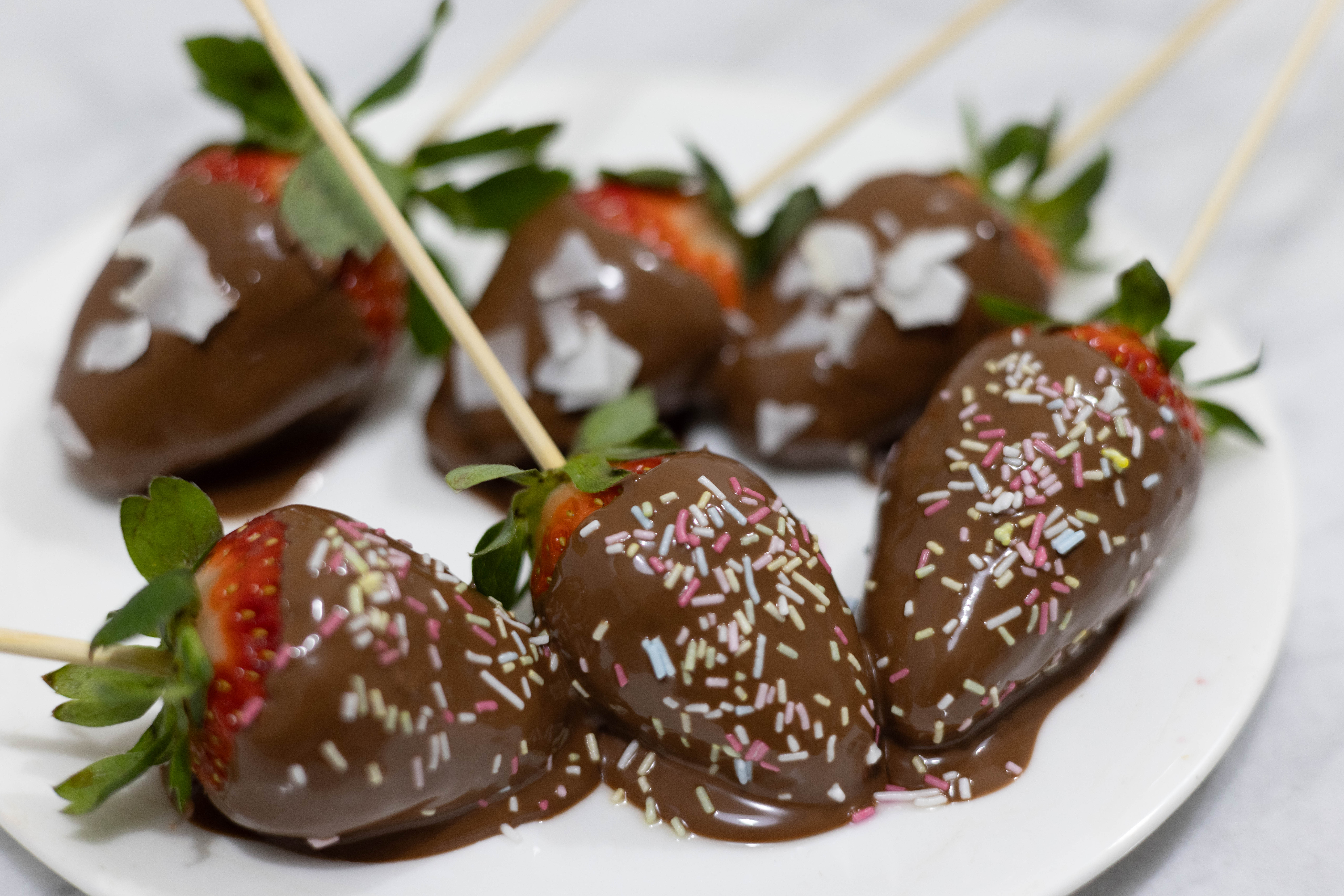 In this picture you can see the six Vegan Chocolate-Covered Strawberries plated. Three are garnished with organic colorful sprinkles and the other three a garnished with shredded coconut.   The six Vegan Chocolate-Covered Strawberries are skewered with a wood stick. Vegan Chocolate-Covered Strawberries are plated on fine bone china porcelain. The whole setting is positioned on a white Carrara marble desk.
