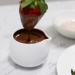 In this picture you can see a jar filled with vegan warm chocolate and a red strawberry is lightly dipped into the chocolate to get it covered. Next to the chocolate jar is a garnish ingredient named organic colorful sprinkles. At the front of the chocolate jar is a white fine bone china porcelain plate filled with a plethora of red organic strawberries. The whole setting is positioned on a white carrara marble desk.