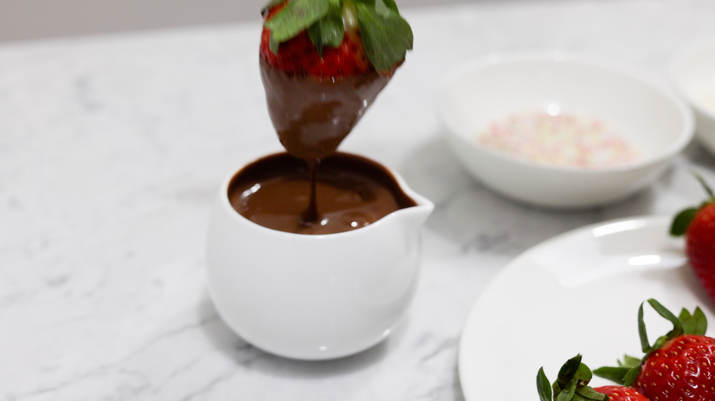 In this picture you can see a fine bone china jar filled with vegan warm chocolate. The red organic strawberry is dipped into the chocolate attached to a wooden skewer. In the background you can see the organic colorful sprinkle garnish. In the front are three red organic strawberries positioned.

The whole setting is positioned on a white Carrara marble desk.
