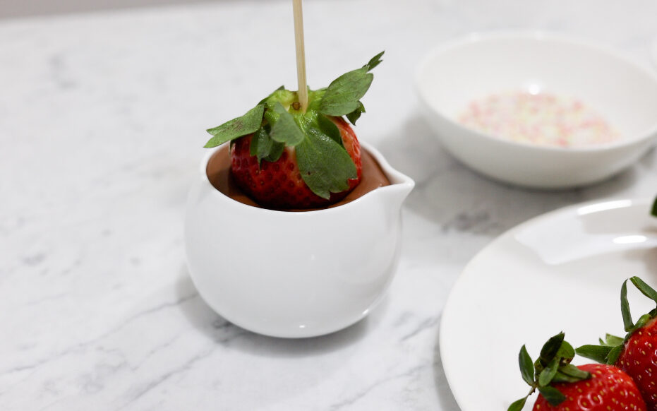 In this picture you can see a fine bone china jar filled with vegan warm chocolate close up. The red organic strawberry is dipped into the chocolate attached to a wooden skewer. In the background you can see the organic colorful sprinkle garnish. In the front are three red organic strawberries positioned.

The whole setting is positioned on a white Carrara marble desk.
