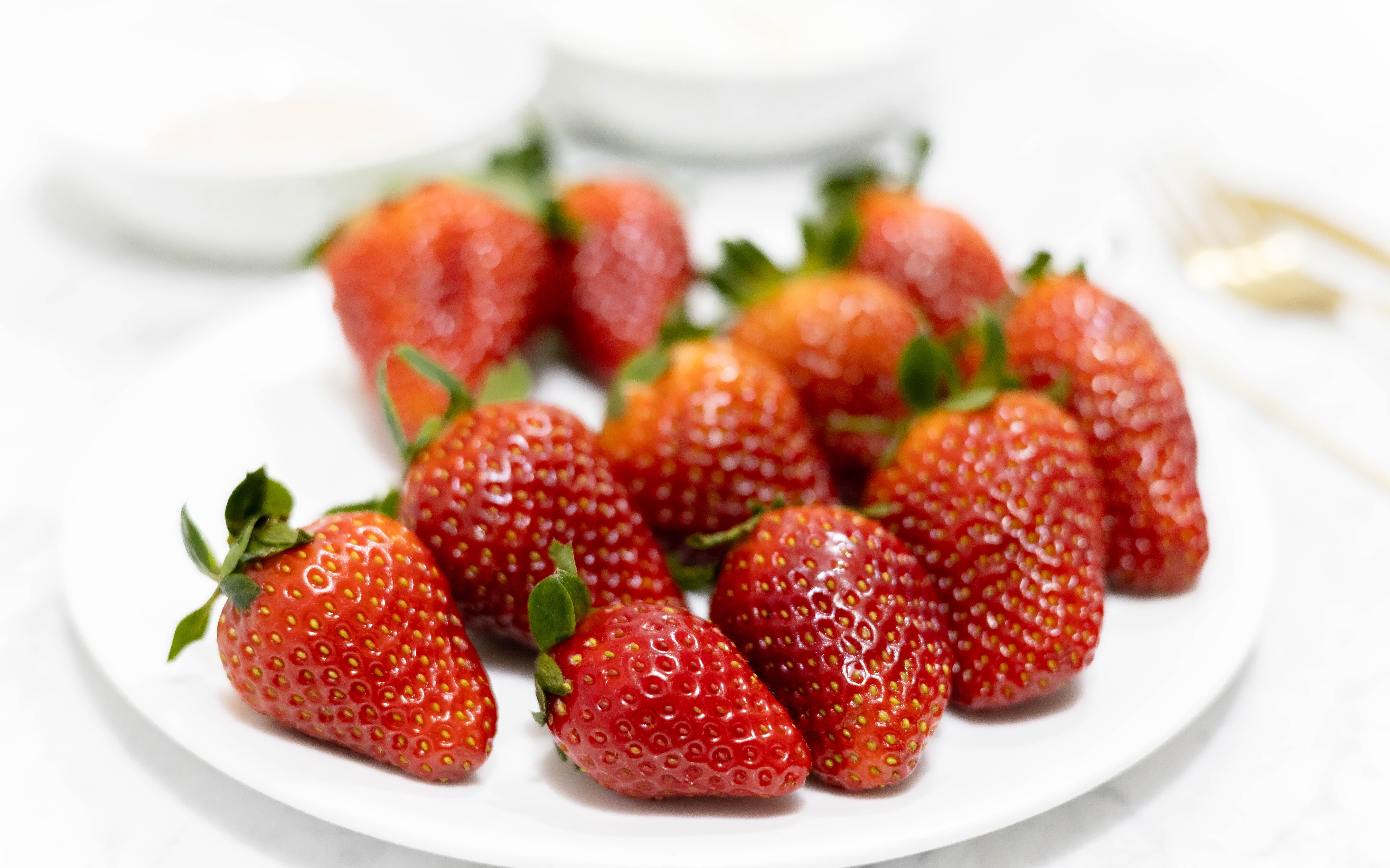 In this picture you can see eight red organic strawberries plated on to a white fine bone china plate. Beside the strawberries is a fine bone china bowl filled with vegan warm chocolate with a golden spoon in it.

The whole setting is positioned on a white Carrara marble desk.
