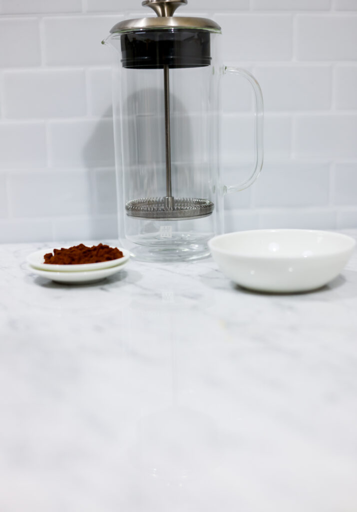 In this picture you can see a French press from the brand Zwilling filled with cold brew coffee. In front of the picture is grounded coffee from the brand the barn coffee. On the right side is a white linen cloth. All is placed on white carrara marble and in the back are white metro tiles.