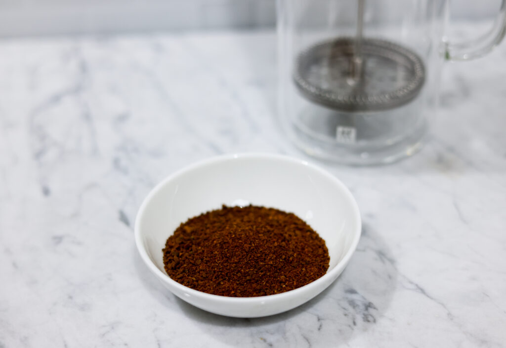 In this picture you can see a French press from the brand Zwilling filled with cold brew coffee. In front of the picture is grounded coffee from the brand the barn coffee. All is placed on white carrara marble and in the back are white metro tiles.