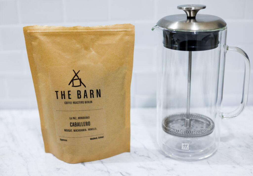 In this picture you can see a French press from the brand Zwilling filled with cold brew coffee. In front of the picture is grounded coffee from the brand the barn coffee. On the right side is a white linen cloth. All is placed on white carrara marble and in the back are white metro tiles.