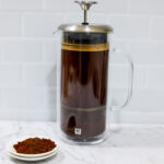 In this picture you can see a French press from the brand Zwilling filled with cold brew coffee. In front of the picture is grounded coffee from the brand the barn coffee. On the right side is a white linen cloth. All objects are placed on white carrara marble and in the back are white metro tiles.