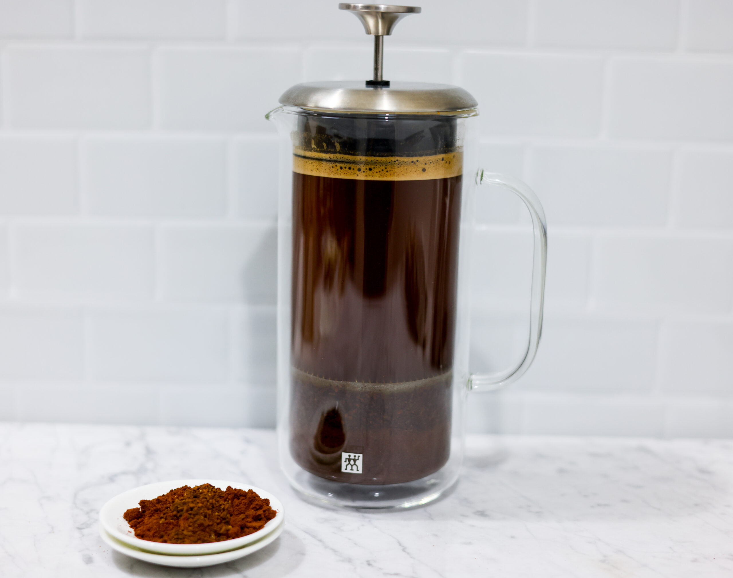 In this picture you can see a French press from the brand Zwilling filled with cold brew coffee. In front of the picture is grounded coffee from the brand the barn coffee. On the right side is a white linen cloth. All objects are placed on white carrara marble and in the back are white metro tiles.