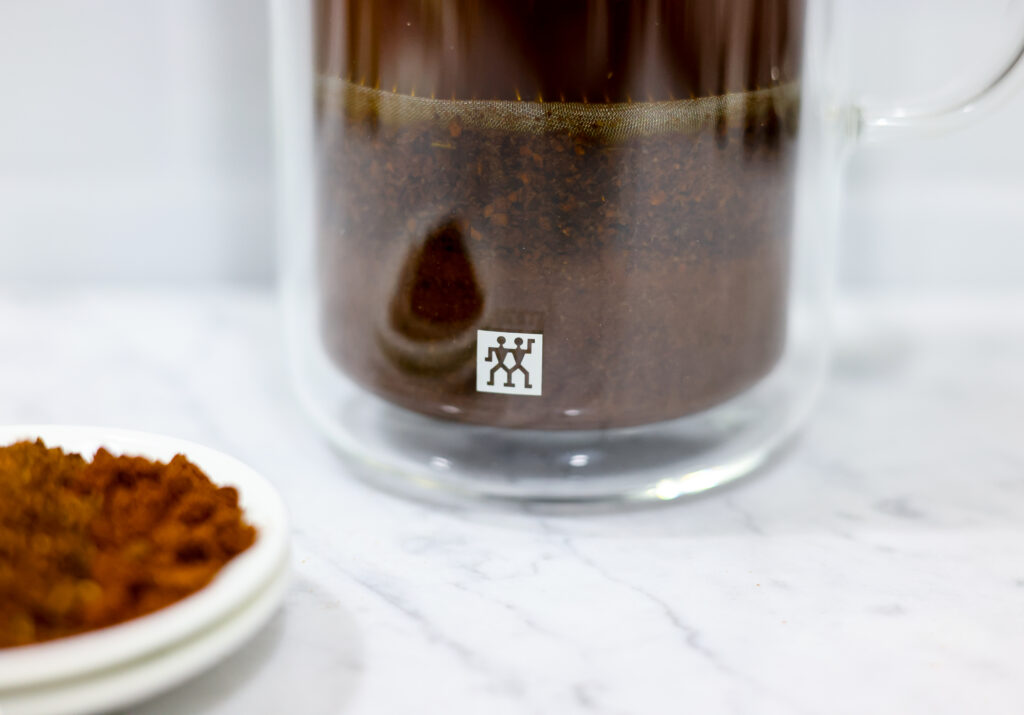 In this picture you can see a French press from the brand Zwilling filled with cold brew coffee. In front of the picture is grounded coffee from the brand the barn coffee. All is placed on white carrara marble and in the back are white metro tiles.