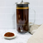 In this picture you can see a French press from the brand Zwilling filled with cold brew coffee. In front of the picture is grounded coffee from the brand the barn coffee. On the right side is a beige linen cloth. All is placed on white carrara marble and in the back are white metro tiles.