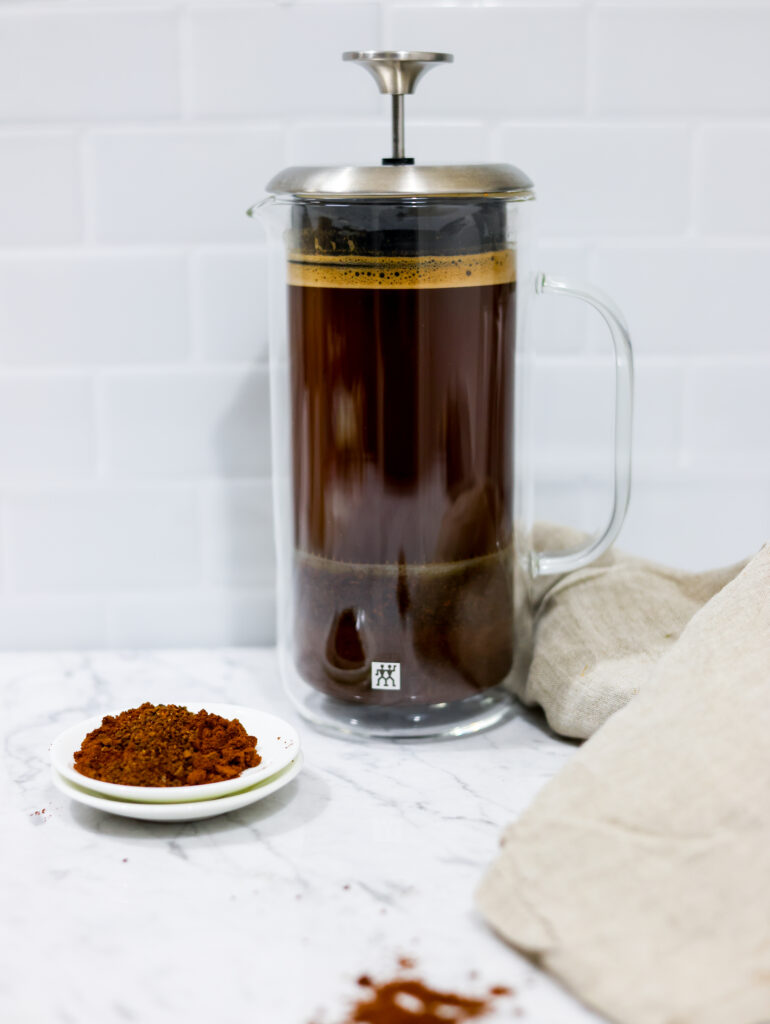 In this picture you can see a French press from the brand Zwilling filled with cold brew coffee. In front of the picture is grounded coffee from the brand the barn coffee. On the right side is a beige linen cloth. All is placed on white carrara marble and in the back are white metro tiles.