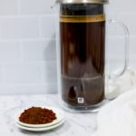 In this picture you can see a French press from the brand Zwilling filled with cold brew coffee. In front of the picture is grounded coffee from the brand the barn coffee. On the right side is a white linen cloth. All is placed on white carrara marble and in the back are white metro tiles.