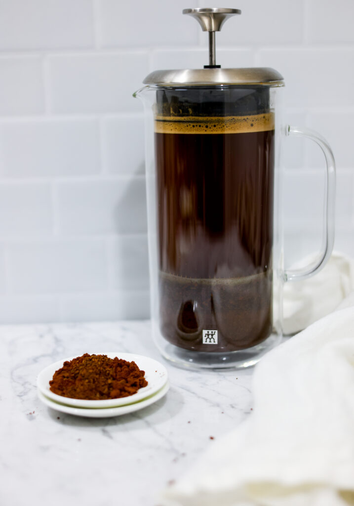 In this picture you can see a French press from the brand Zwilling filled with cold brew coffee. In front of the picture is grounded coffee from the brand the barn coffee. On the right side is a white linen cloth. All is placed on white carrara marble and in the back are white metro tiles.