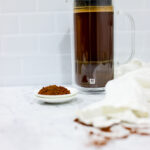 In this picture you can see a French press from the brand Zwilling filled with cold brew coffee. In front of the picture is grounded coffee from the brand the barn coffee. On the right side is a white linen cloth. All is placed on white carrara marble and in the back are white metro tiles.