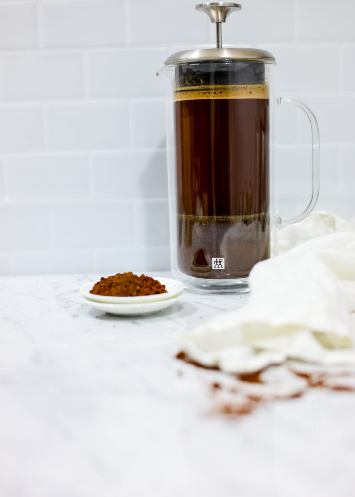 In this picture you can see a French press from the brand Zwilling filled with cold brew coffee. In front of the picture is grounded coffee from the brand the barn coffee. On the right side is a white linen cloth. All is placed on white carrara marble and in the back are white metro tiles.