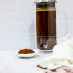 In this picture you can see a French press from the brand Zwilling filled with cold brew coffee. In front of the picture is grounded coffee from the brand the barn coffee. On the right side is a white linen cloth. All objects are placed on white carrara marble and in the back are white metro tiles.