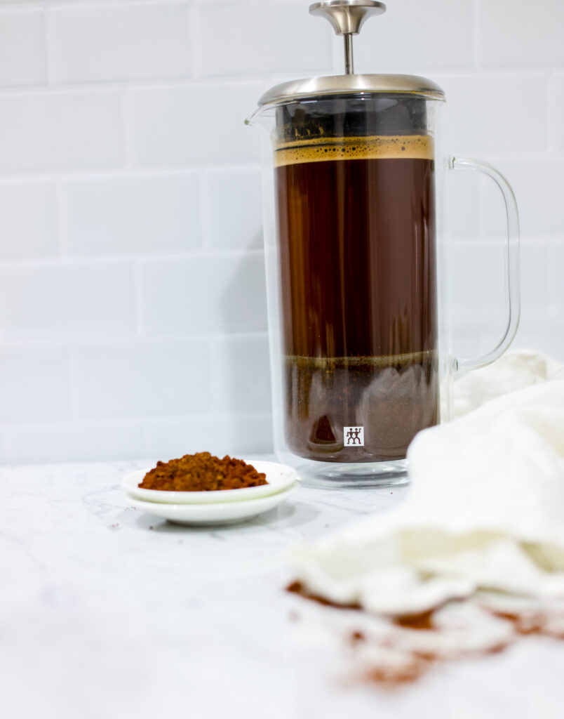 In this picture you can see a French press from the brand Zwilling filled with cold brew coffee. In front of the picture is grounded coffee from the brand the barn coffee. On the right side is a white linen cloth. All objects are placed on white carrara marble and in the back are white metro tiles.