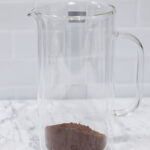 In this picture you can see a French press from the brand Zwilling filled with coarsely grounded the barn coffee beans. All objects are placed on white carrara marble and in the back are white metro tiles.