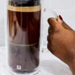 In this picture you can see a French press from the brand Zwilling filled with coarsely grounded the barn coffee beans. The coffee beans are being pressed down. All objects are placed on white carrara marble and in the back are white metro tiles.