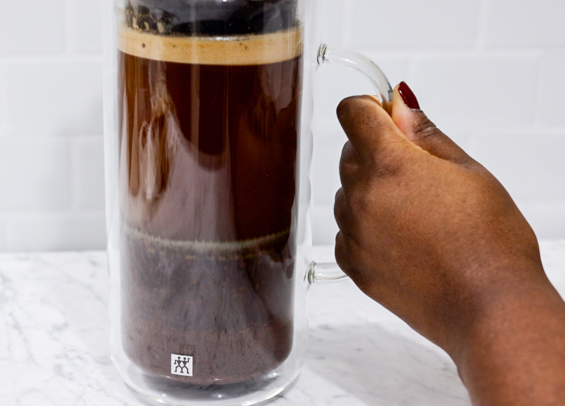 In this picture you can see a French press from the brand Zwilling filled with coarsely grounded the barn coffee beans. The coffee beans are being pressed down. All objects are placed on white carrara marble and in the back are white metro tiles.