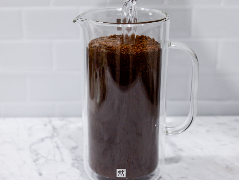 In this picture you can see a French press from the brand Zwilling filled with coarsely grounded the barn coffee beans. All objects are placed on white carrara marble and in the back are white metro tiles.