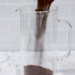 In this picture you can see a French press from the brand Zwilling filled with coarsely grounded the barn coffee beans. All objects are placed on white carrara marble and in the back are white metro tiles.