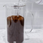 In this picture you can see a French press from the brand Zwilling filled with coarsely grounded the barn coffee beans and filtered water. All objects are placed on white carrara marble and in the back are white metro tiles.