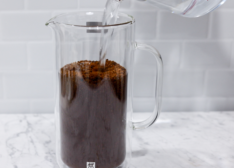 In this picture you can see a French press from the brand Zwilling filled with coarsely grounded the barn coffee beans and filtered water. All objects are placed on white carrara marble and in the back are white metro tiles.