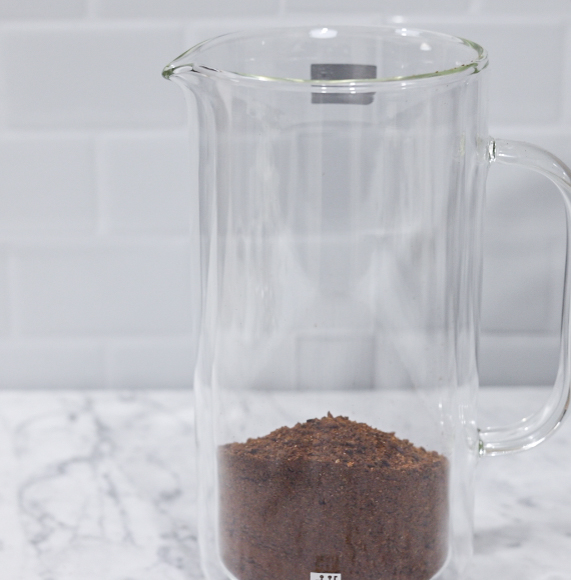 In this picture you can see a French press from the brand Zwilling filled with coarsely grounded the barn coffee beans. All objects are placed on white carrara marble and in the back are white metro tiles.