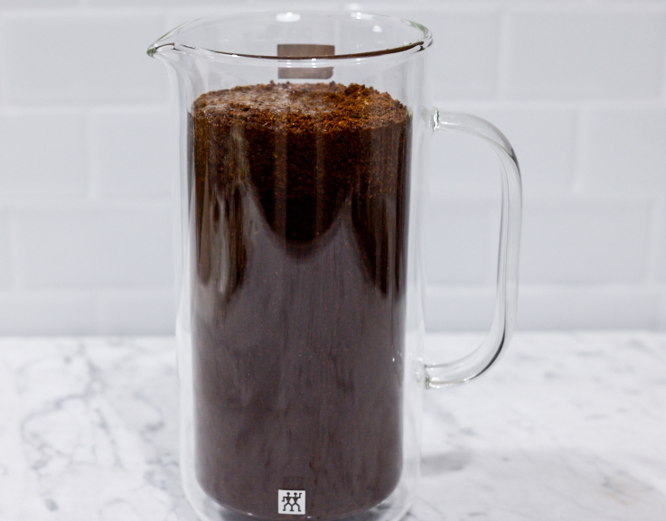 In this picture you can see a French press from the brand Zwilling filled with coarsely grounded the barn coffee beans. All objects are placed on white carrara marble and in the back are white metro tiles.