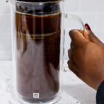 In this picture you can see a French press from the brand Zwilling filled with coarsely grounded the barn coffee beans. The coffee beans are being pressed down. All objects are placed on white carrara marble and in the back are white metro tiles.