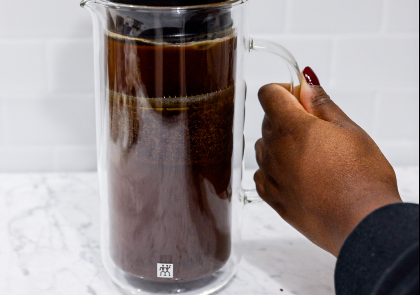 In this picture you can see a French press from the brand Zwilling filled with coarsely grounded the barn coffee beans. The coffee beans are being pressed down. All objects are placed on white carrara marble and in the back are white metro tiles.