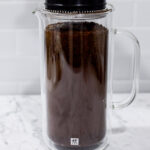 In this picture you can see a French press from the brand Zwilling filled with coarsely grounded the barn coffee beans. All objects are placed on white carrara marble and in the back are white metro tiles.