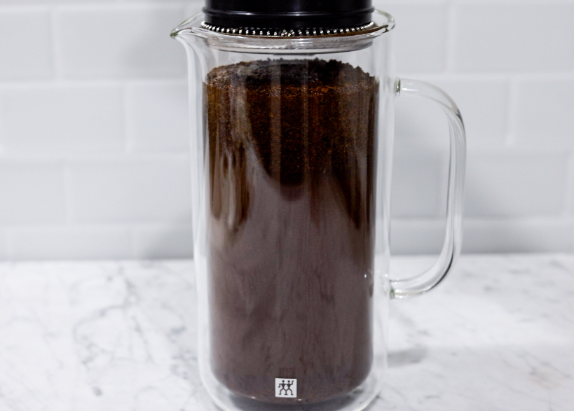 In this picture you can see a French press from the brand Zwilling filled with coarsely grounded the barn coffee beans. All objects are placed on white carrara marble and in the back are white metro tiles.