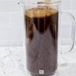 In this picture you can see a French press from the brand Zwilling filled with coarsely grounded the barn coffee beans. The coffee is being stirred by a glass straw. All objects are placed on white carrara marble and in the back are white metro tiles.