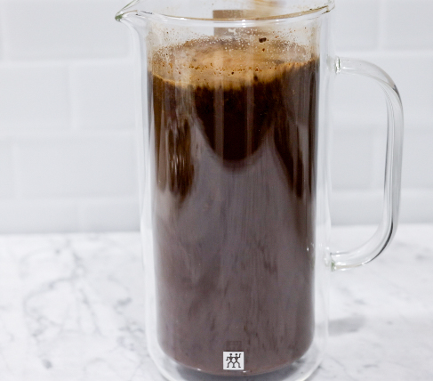 In this picture you can see a French press from the brand Zwilling filled with coarsely grounded the barn coffee beans. The coffee is being stirred by a glass straw. All objects are placed on white carrara marble and in the back are white metro tiles.